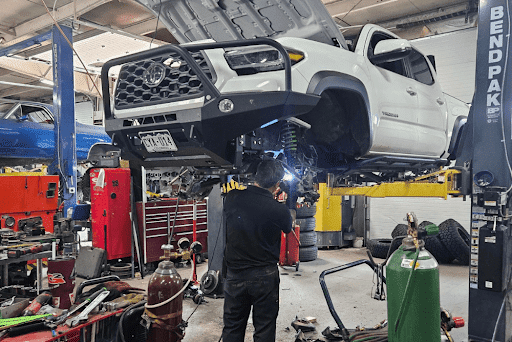 Is your off-road vehicle hunting season ready in Grand Junction, CO? Image of Barney Brothers Off Road and Repair mechanic performing suspension repairs on a white Toyota Tacoma in shop with other tools and vehicles around.