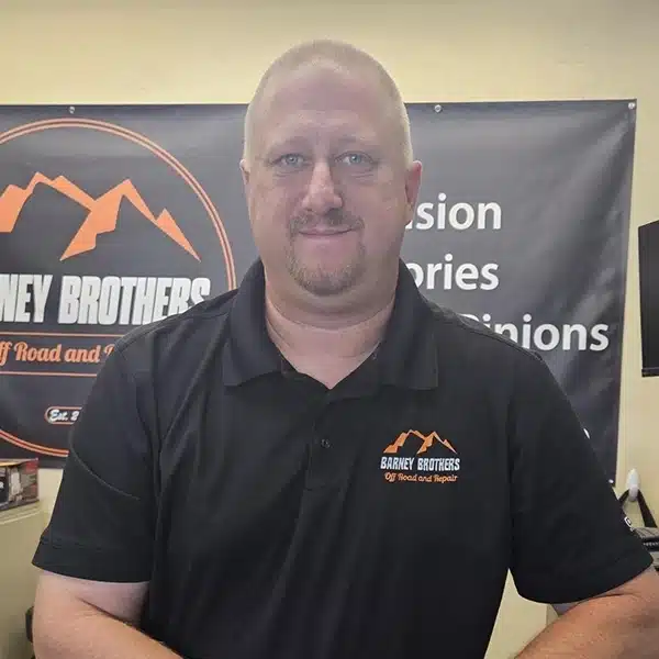 Erik Rudolph standing behind the service counter at Barney Brothers Off Road and Repair, wearing a branded black polo in a professional automotive office featuring suspension and fabrication signage.